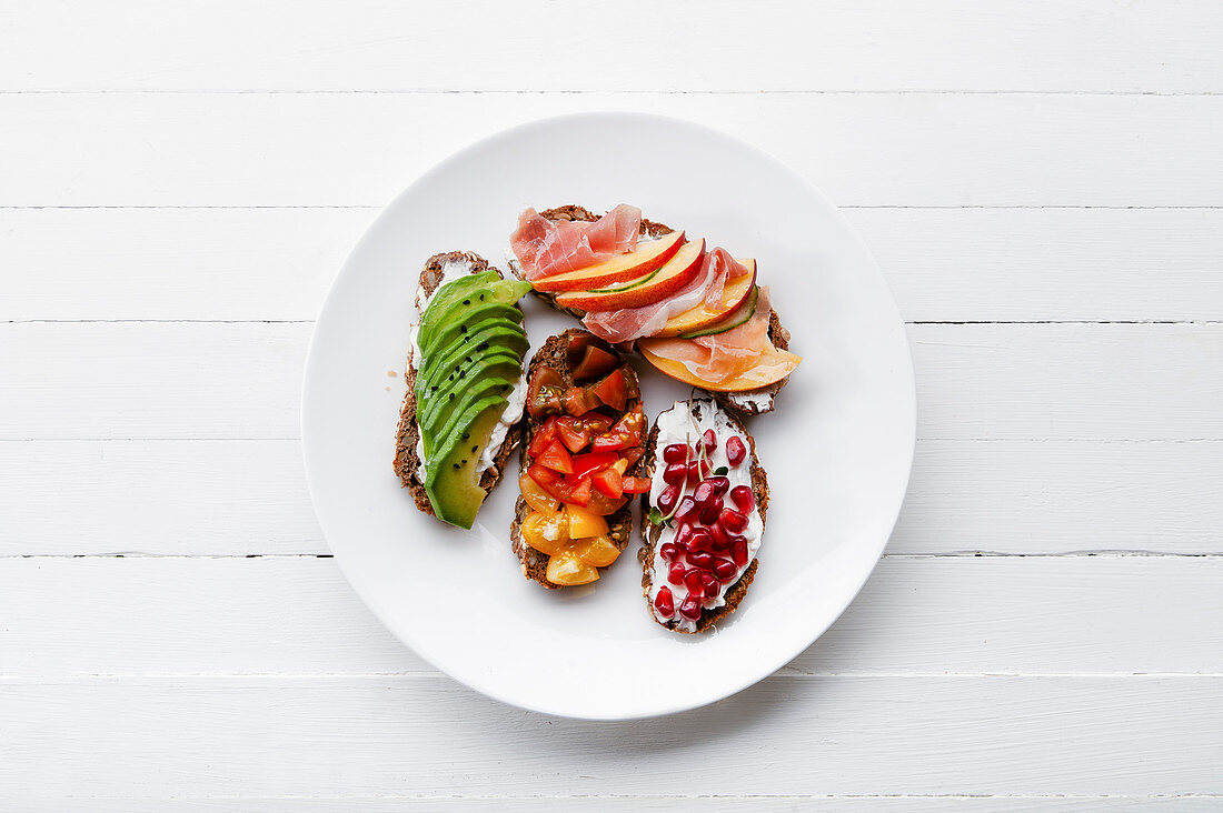 Variety of bruschettas with avecado, prosciutto, tomatoes and pomegranate seeds on white plate