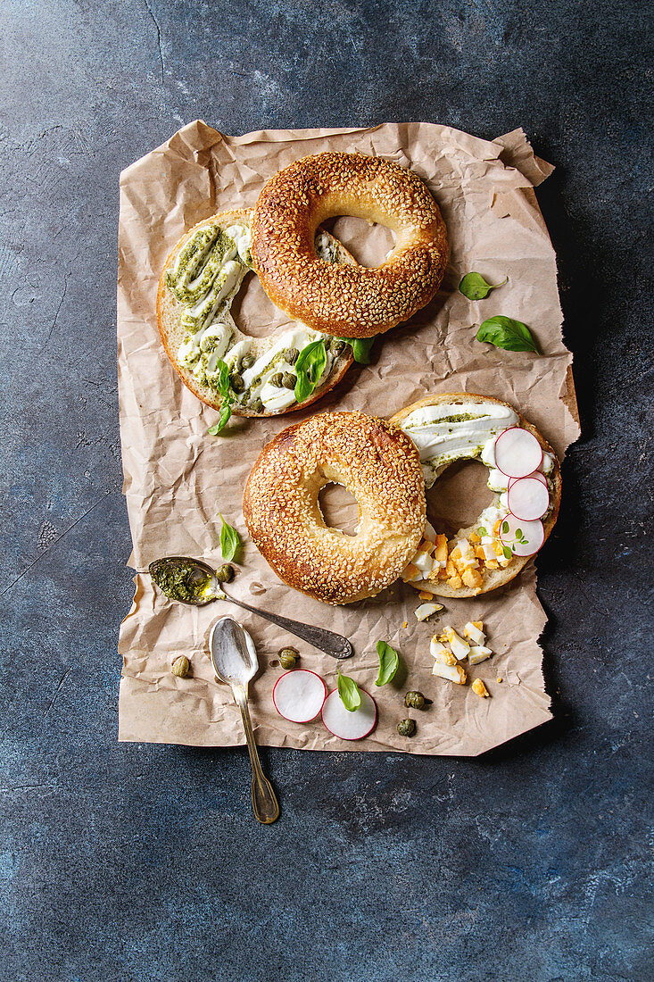 Variety of homemade bagels with sesame seeds, cream cheese, pesto sauce, eggs, radish, herbs