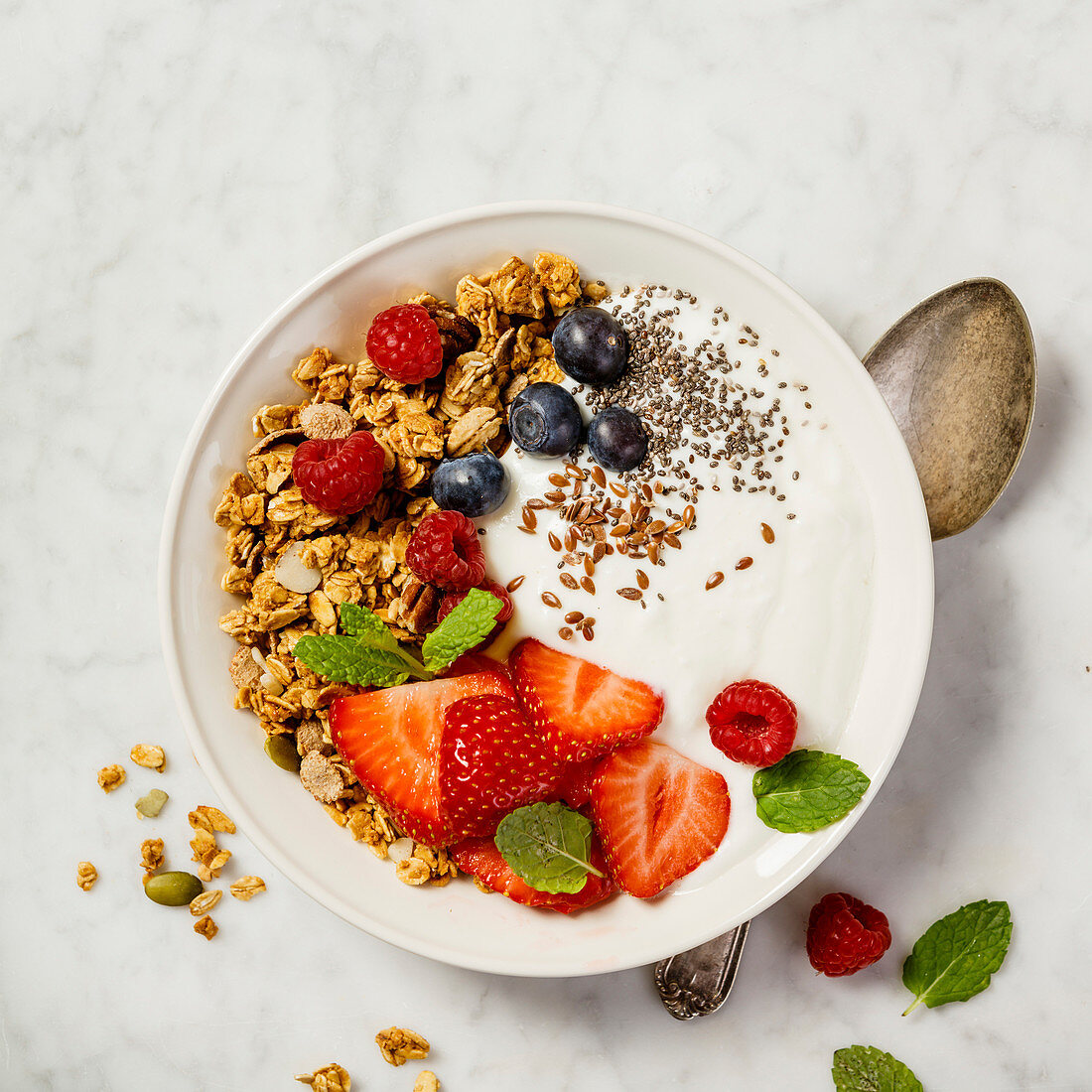 Bowl of homemade granola with yogurt and fresh berries on white marble background