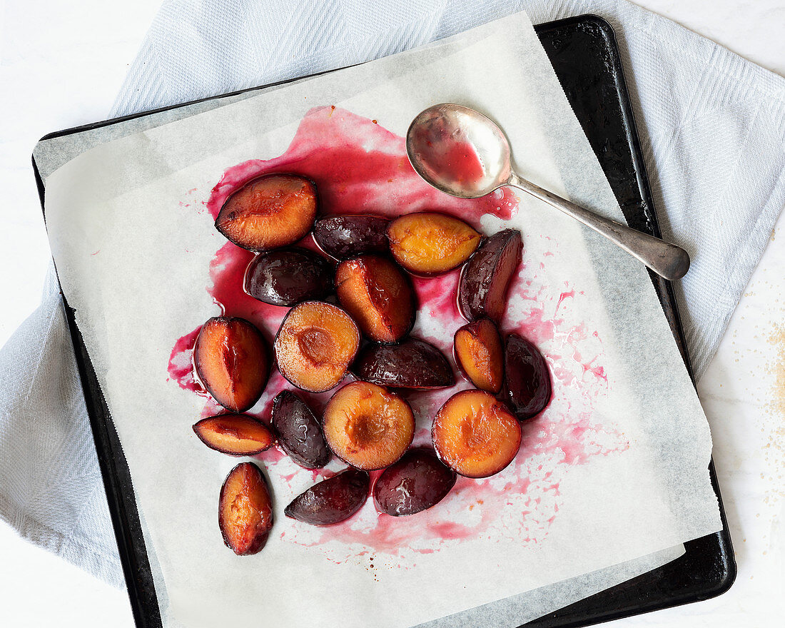 Roasted red plums in syrup with a spoon on a baking tray