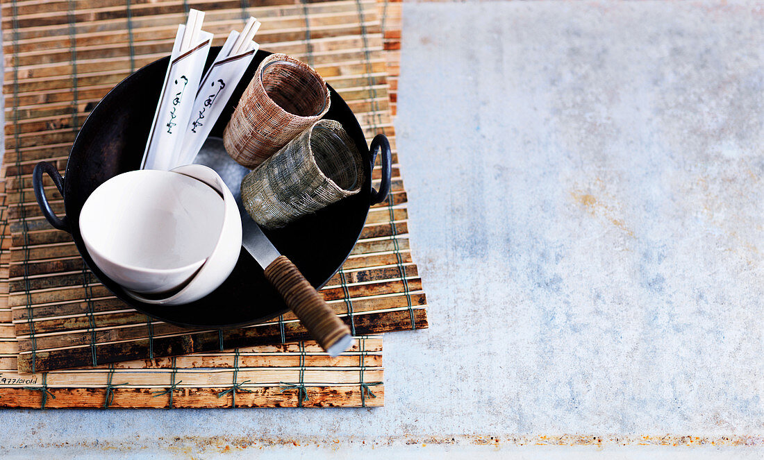 A wok, small bowls, serving spoons, glasses and chopsticks on a bamboo mat