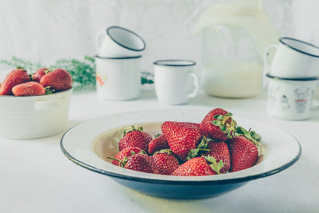 A plate with fresh strawberries, mugs, milk in a glass jar on a white background