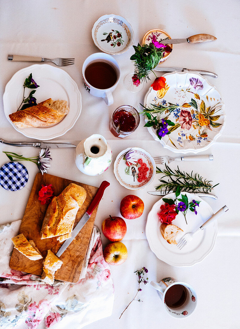 A table with a white cloth with French bread, marmelade, apples and tea