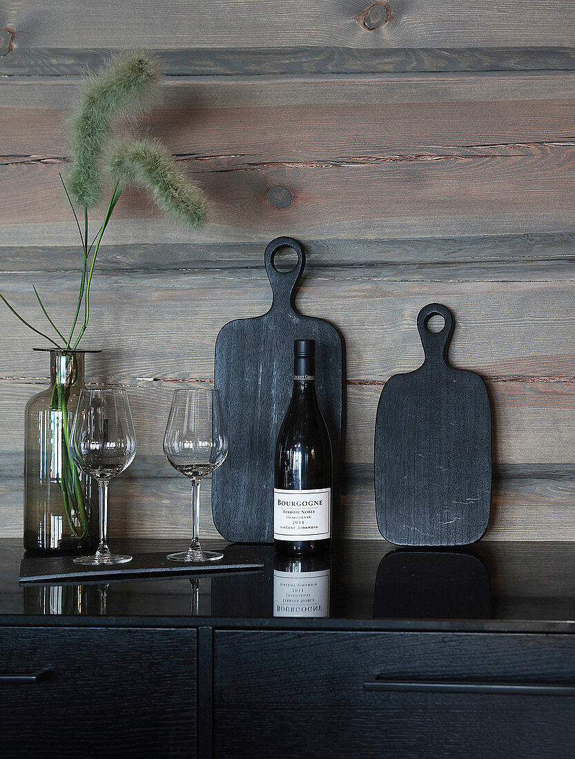 Schwarze Schneidebretter, Weinflasche und Gläser vor einer Holzwand