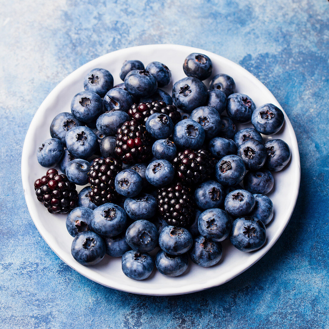 Blueberry and blackberry berries on a white plate on blue stone background