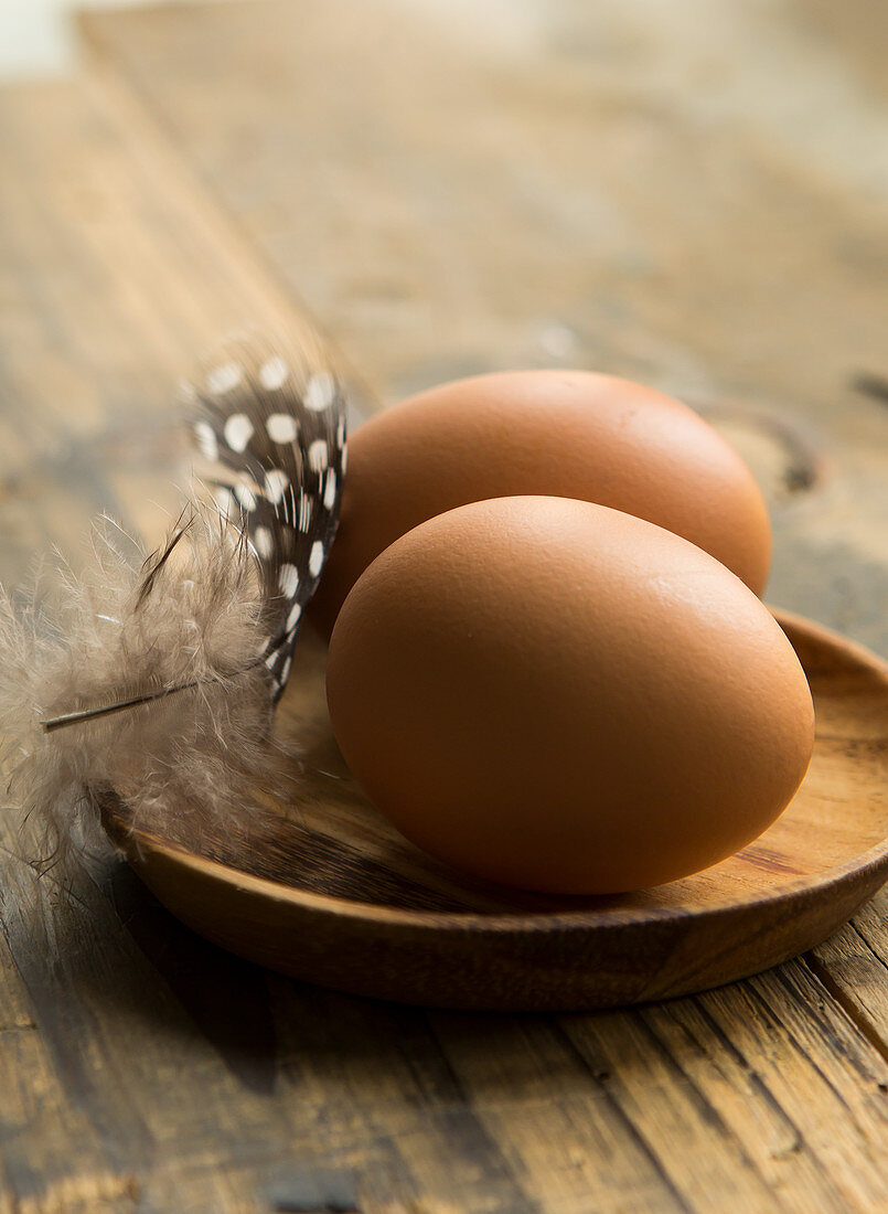 Two fresh eggs in a wooden dish and a spotty feather on a rustic wooden table