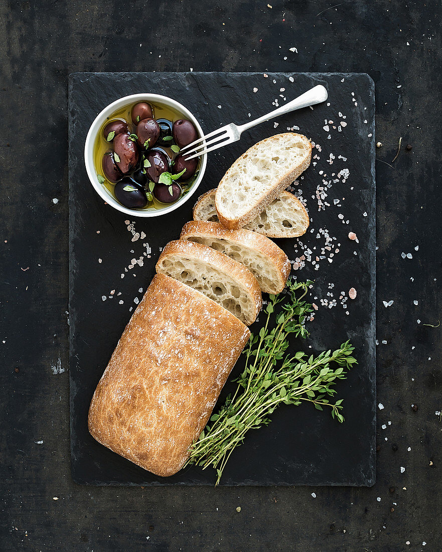 Italian ciabatta bread cut in slices on wooden chopping board with herbs, garlic and olives