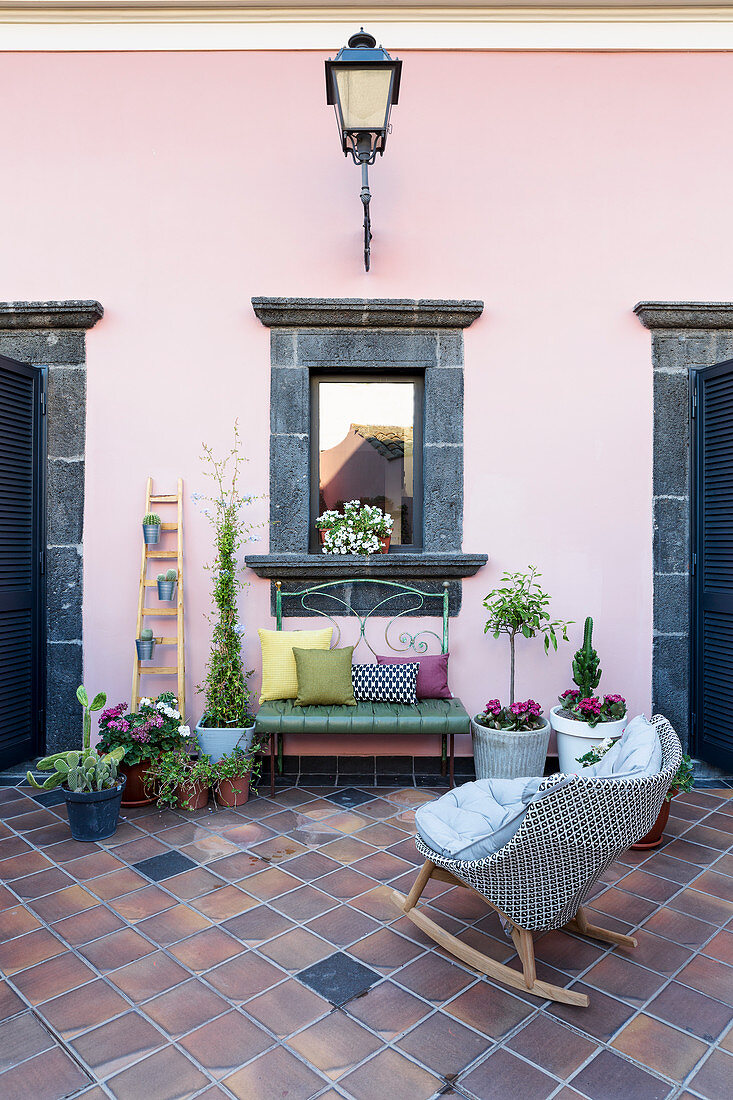 Schaukelstuhl, Grünpflanzen, Bank und verspiegeltes Fenster mit Rahmen aus Lavastein an rosa Wand im Innenhof