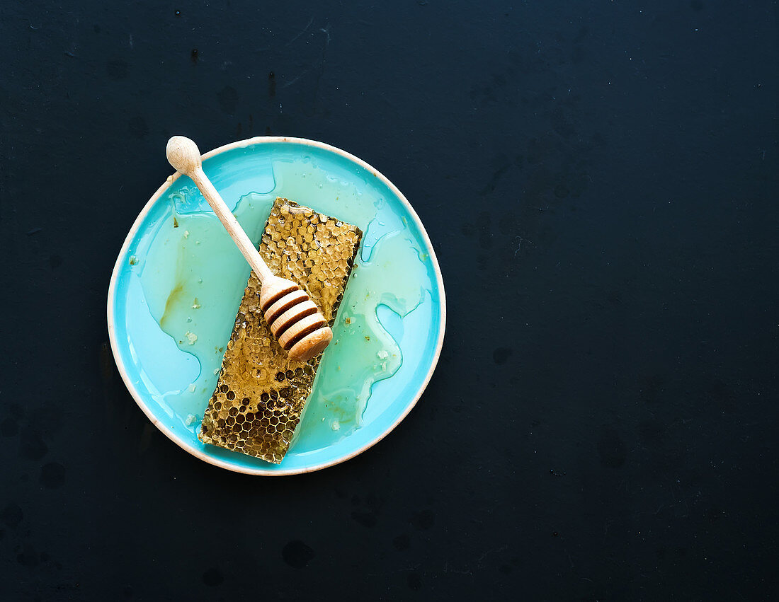 Honeycomb with honey dipper on blue ceramic plate over black background, top view copy space