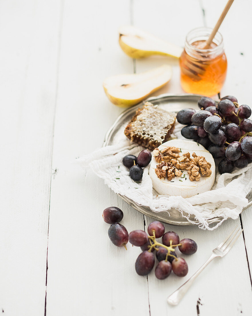 Camembert cheese with grape, walnuts, pear and honey on vintage metal plate over white rustic wood backdrop
