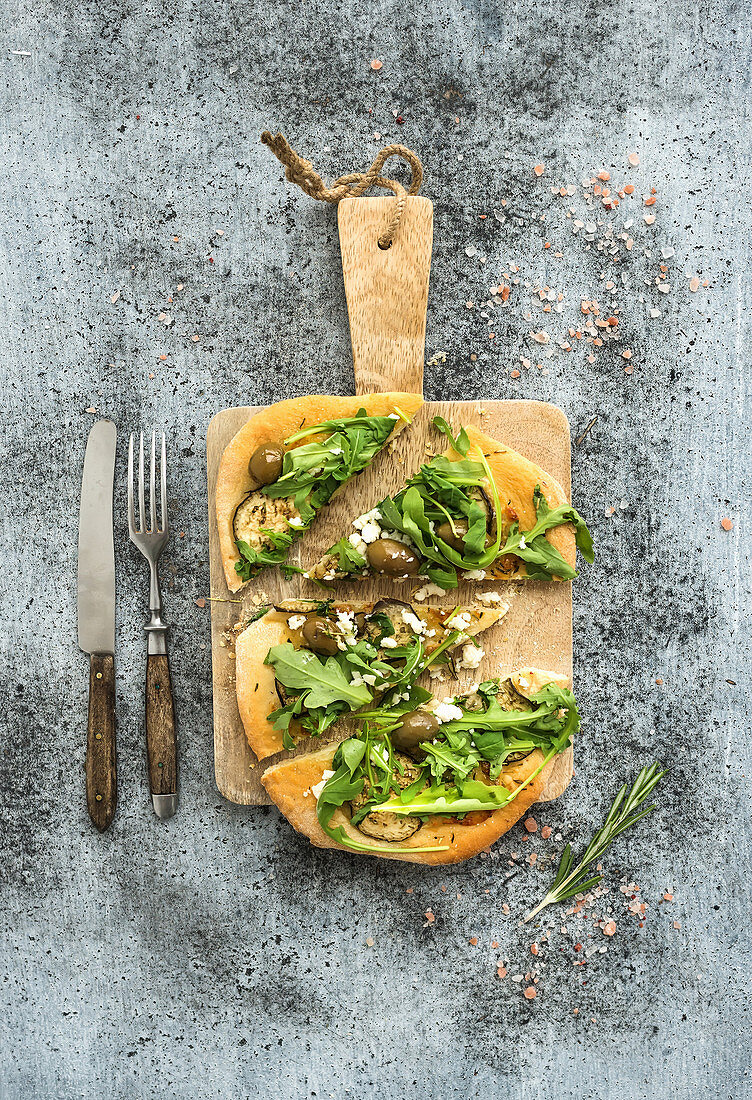 Rustic homemade pizza with eggpant, cheese, olives and arugula on rustic wooden serving board over grunge backdrop