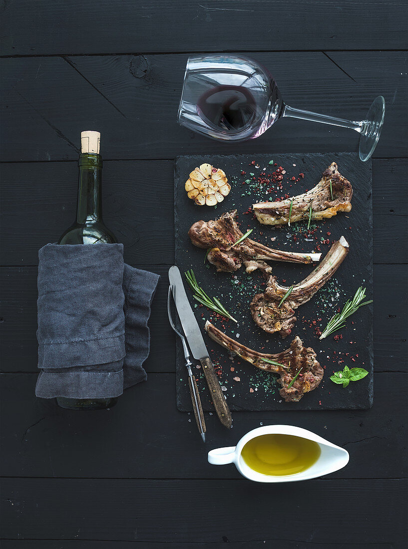 Grilled lamb chops. Rack of Lamb with garlic, rosemary and spices on slate tray, wine glass, oil in a saucer and bottle over black wood background