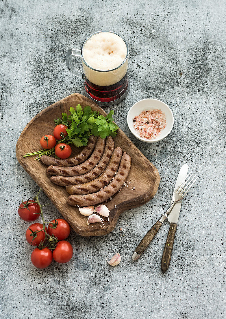 Grilled sausages with vegetables on rustic serving board and mug of light beer over grunge backdrop