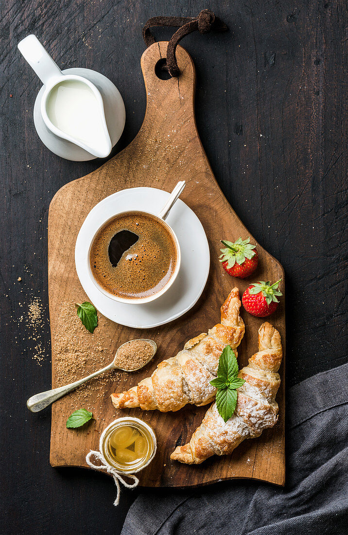 Freshly baked croissants with strawberries, honey, cup of black coffee, pitcher with milk and spoon with brown sugar