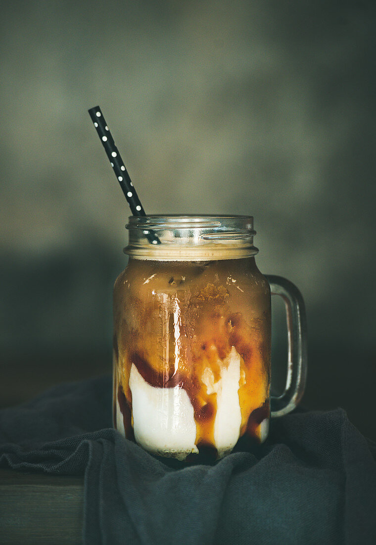 Iced caramel macciato coffee with milk in glass jar with straw on dark rustic wooden table