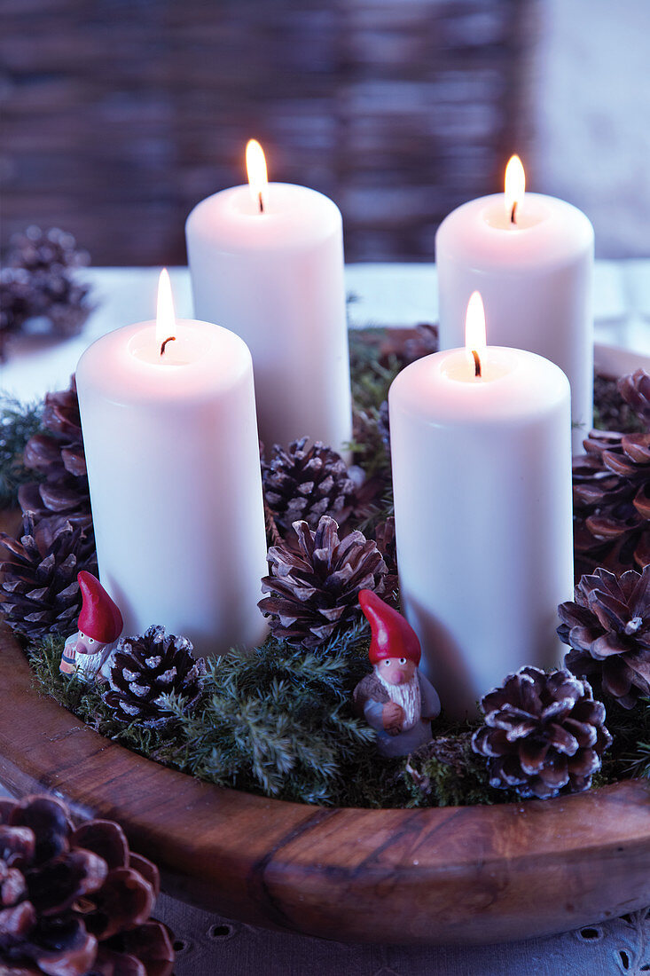 Four pillar candles in wooden bowl festively decorated with moss, pine cones and gnome figurines