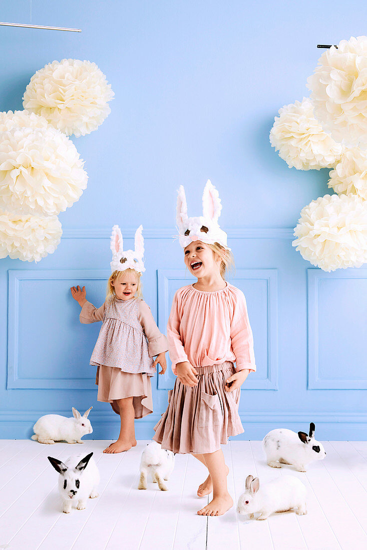 Two girls with DIY rabbit masks and lively bunnies in the room with a light blue wall