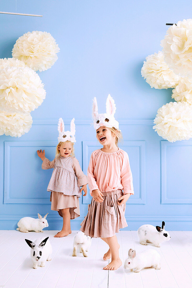 Zwei Mädchen mit DIY-Hasen-Masken und lebendigen Häschen im Raum mit hellblauer Wand