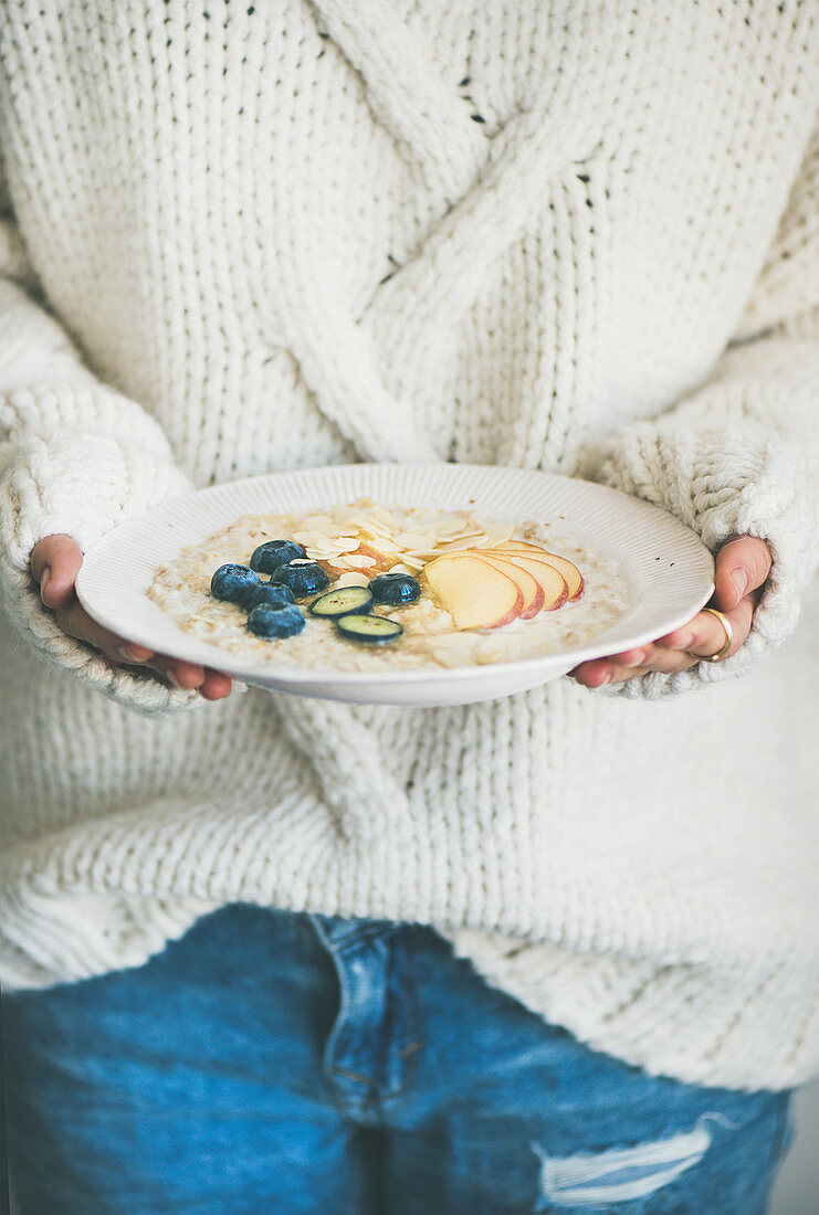 Woman in woolen sweater and jeans holding plate of vegan almond milk oatmeal porridge with berries, fruit and almonds