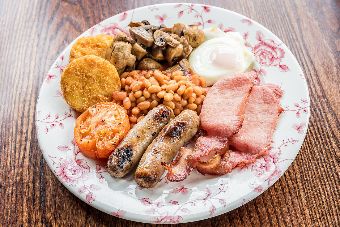 Traditional english breakfast with bacon, sausages, grilled tomato and mushrooms, baked beans, potato hash browns cakes and egg on a wooden table