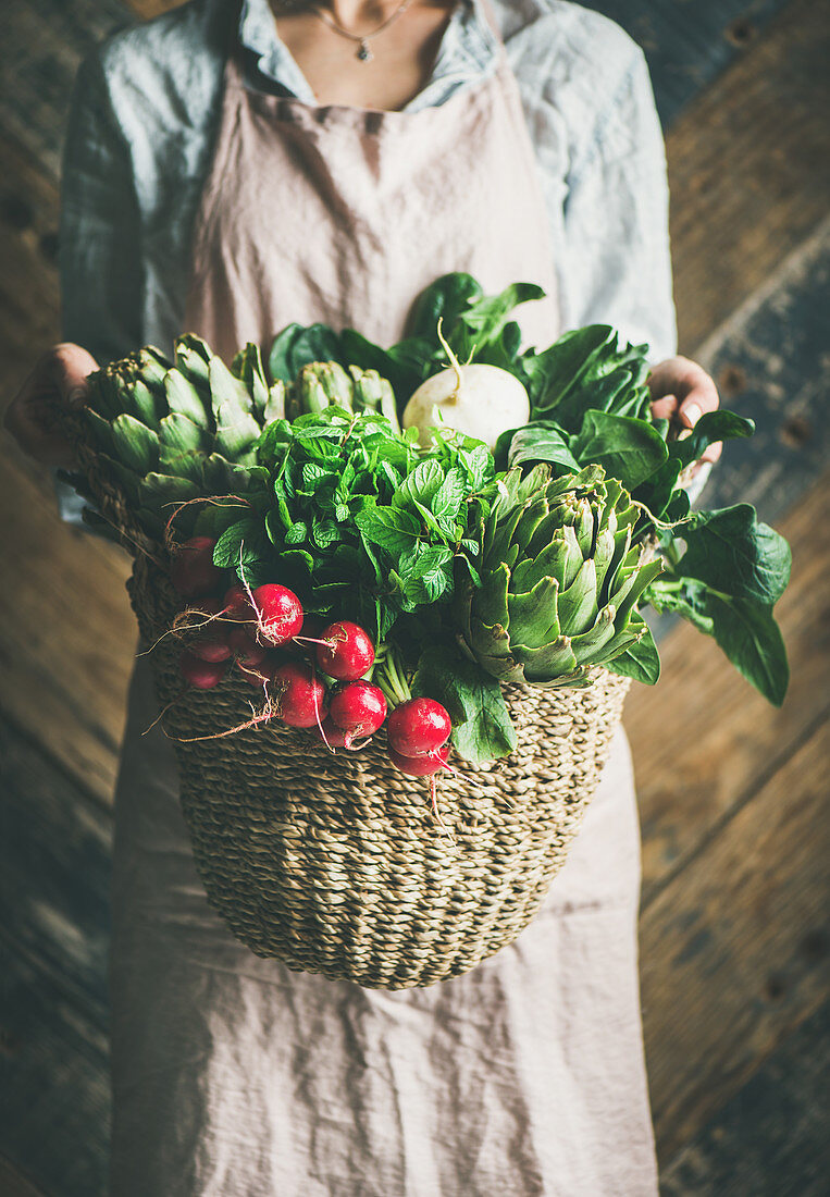 Female farmer in linen apron holding basket of fresh garden vegetables and greens in her hands