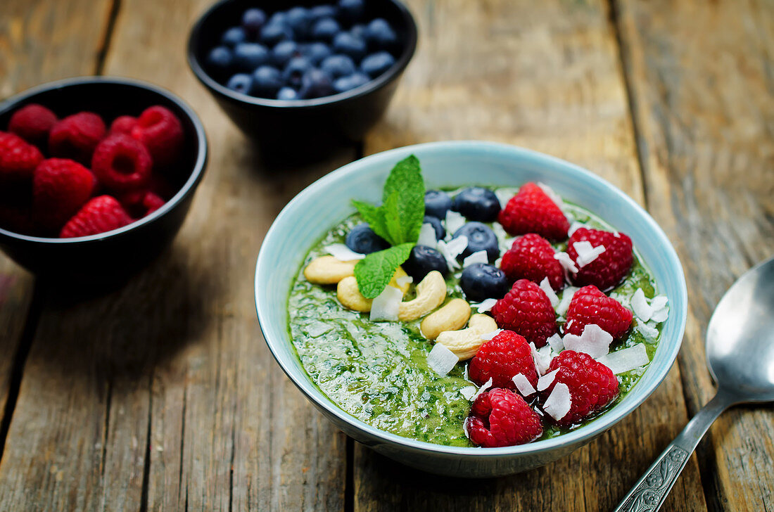 Breakfast bowl mit Spinatsmoothie, Himbeeren, Cashewkernen, Blaubeeren und Kokosflocken