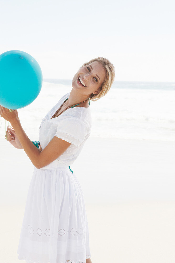 A blonde woman on a beach with a blue balloon wearing a white bolero and an embroidered skirt