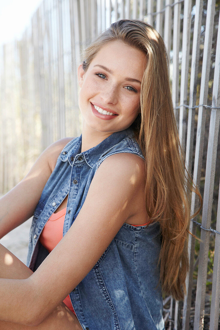 A young blonde woman sitting against a fence wearing a denim jacket