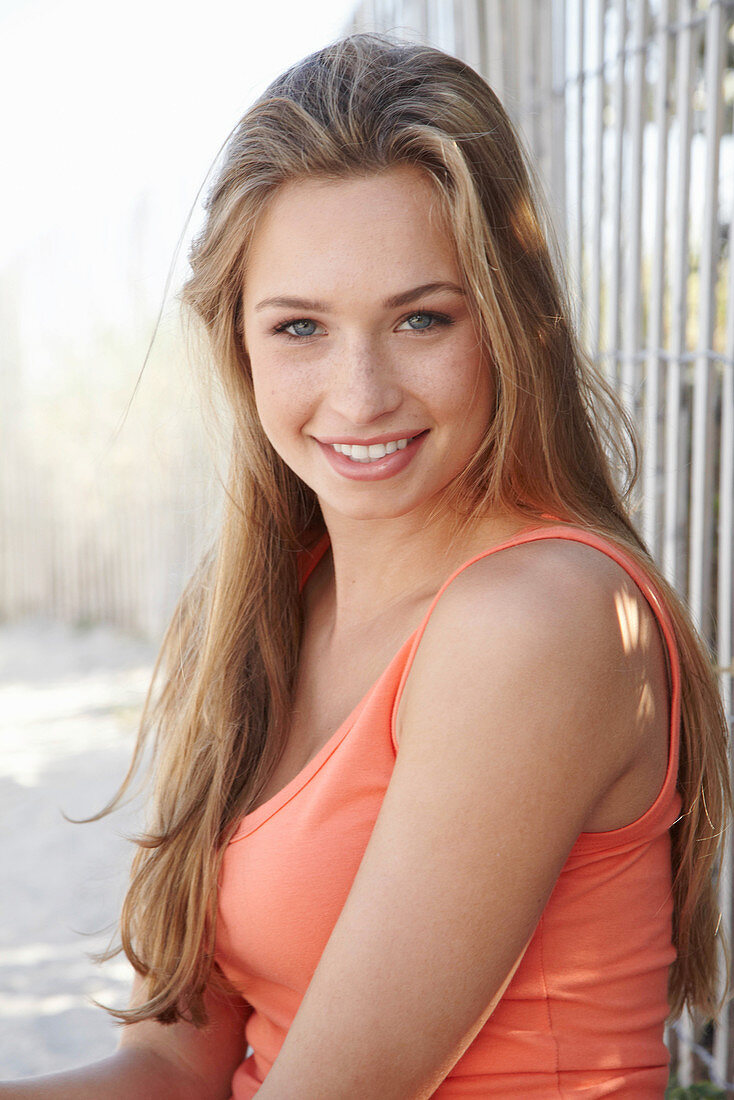 A young blonde woman sitting against a fence wearing an orange top