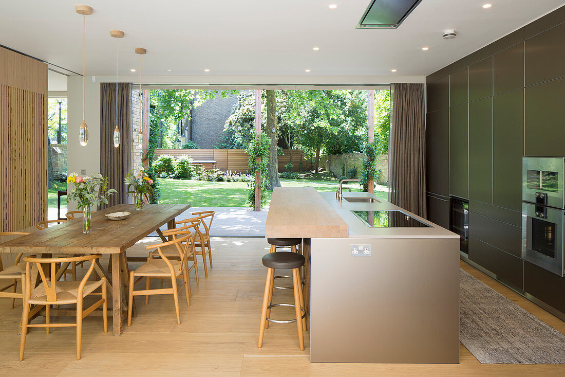 Designer-Einbauküche mit Kücheninsel und rustikaler Esstisch mit Klassikerstühlen, im Hintergrund Gartenblick