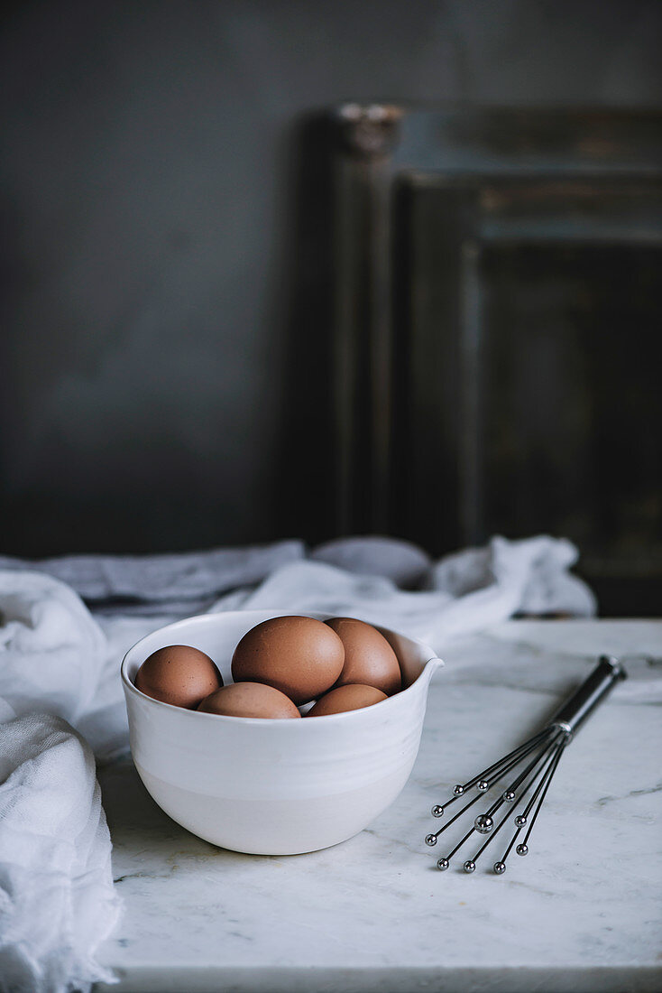 Eier in weisser Schale auf Marmortisch in Küche, daneben Schneebesen