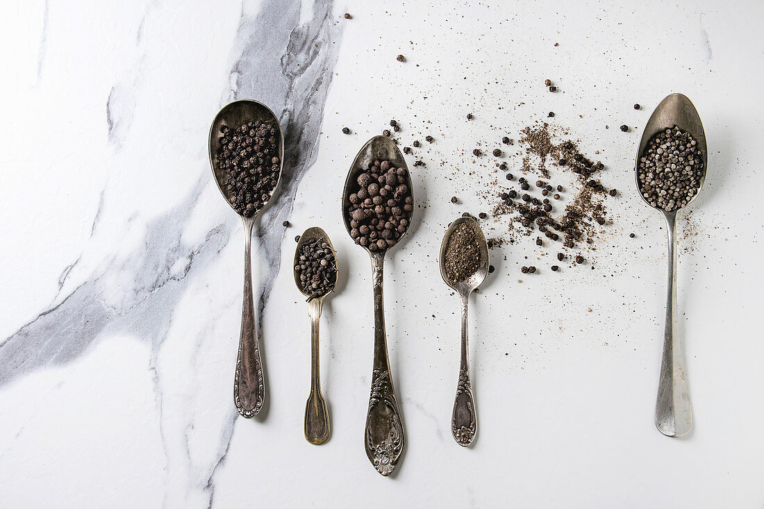 Variety of different black peppers allspice, pimento, long pepper, monks pepper, peppercorns and ground powder in vintage spoons over white marble texture background
