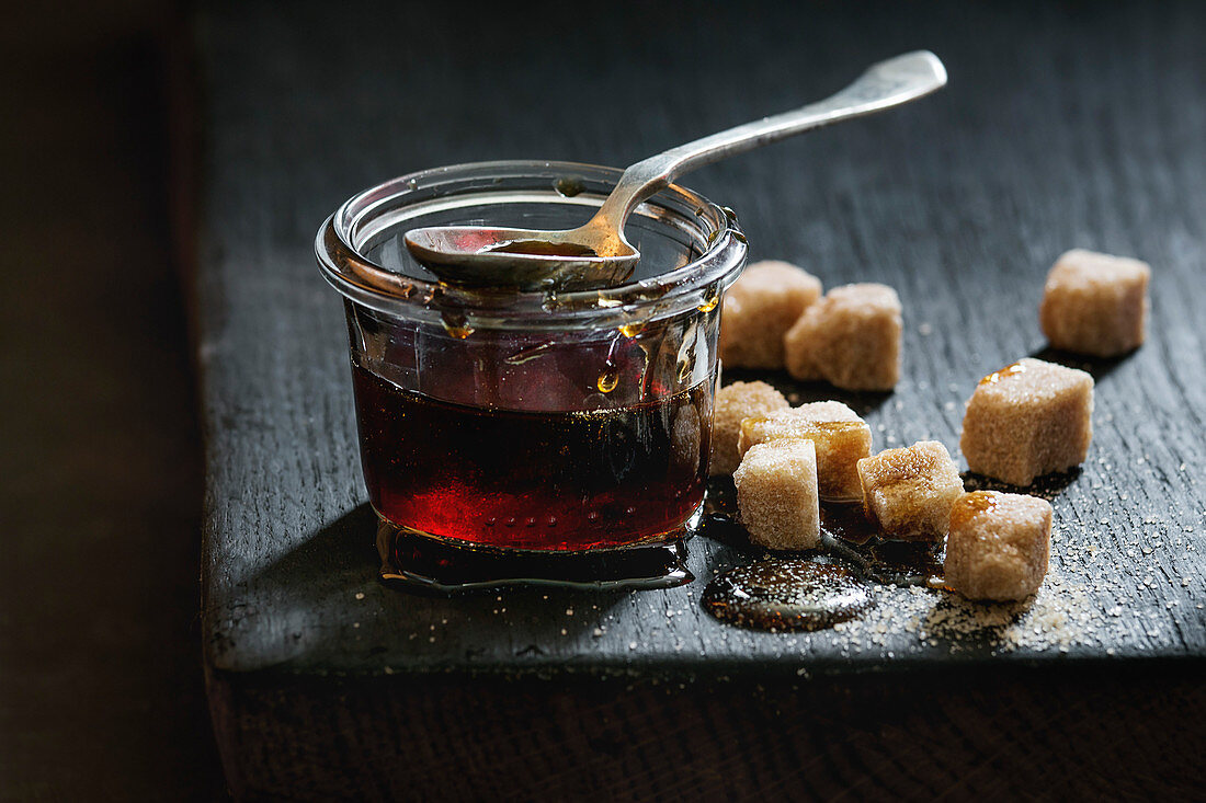 Homemade liquid transparent brown sugar caramel in glass jar standing on black wooden board with spoon and can sugar cubes