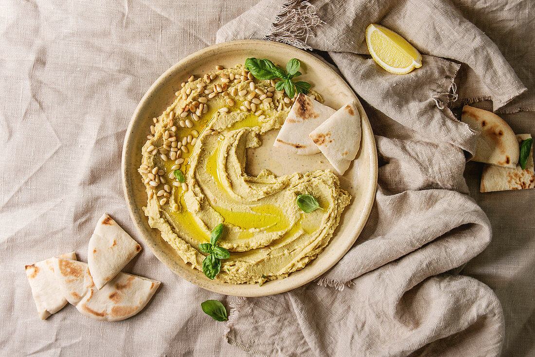 Homemade traditional spread hummus with pine nuts, olive oil, basil served on ceramic plate with pita bread over linen cloth background