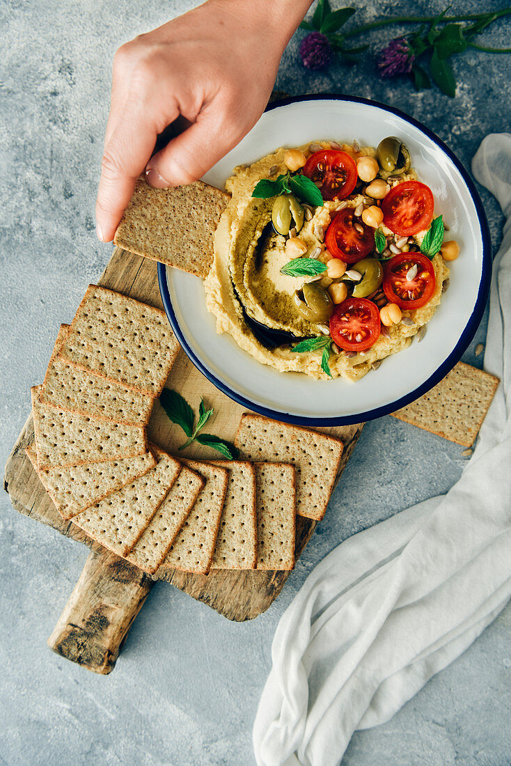 A woman dipping a cracker into a bowl of hummus without tahini topped with cherry tomatoes and green olives