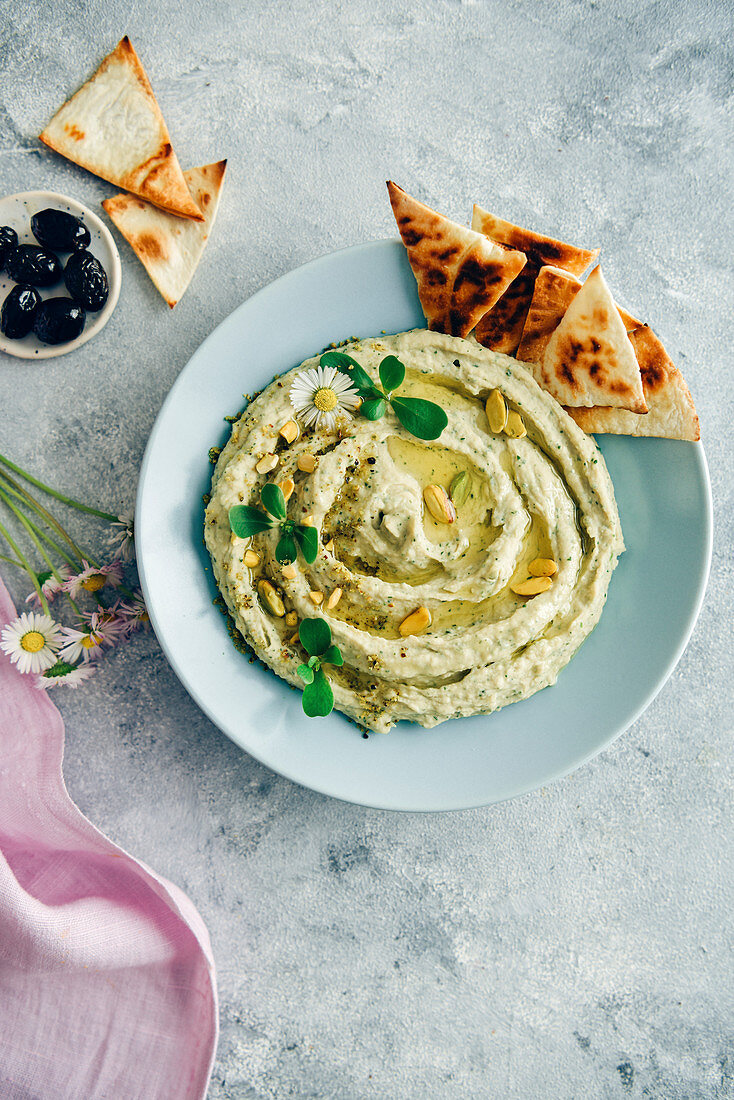 Creamy white bean hummus topped with herbs, olive oil and pistachios served in a light blue bowl with pita chips on the side