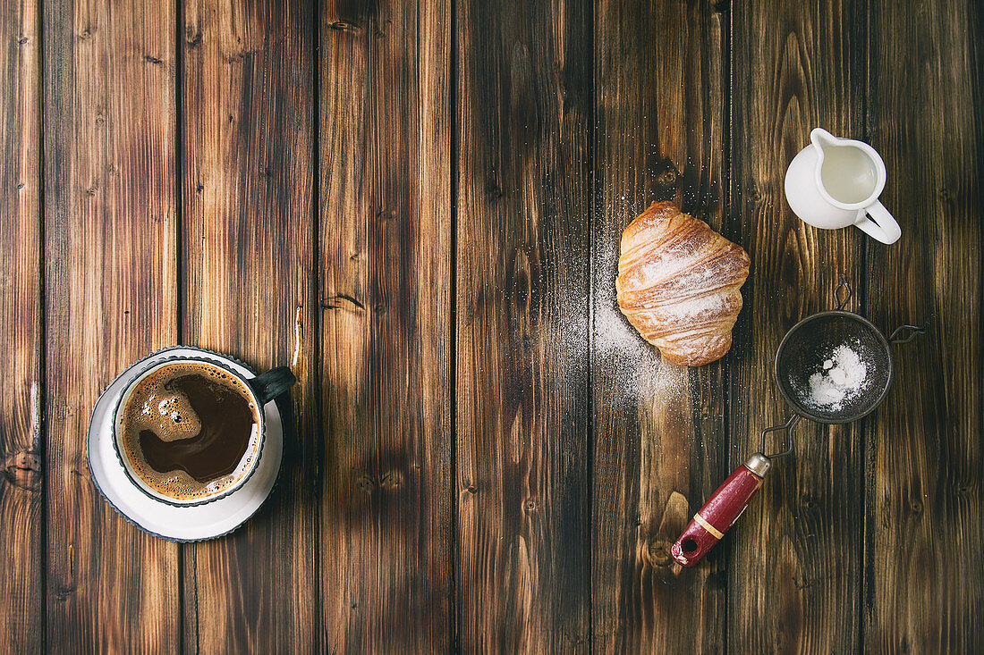 Homemade croissant with sugar powder, cup of coffee, jug of milk, vintage sieve over wooden plank background