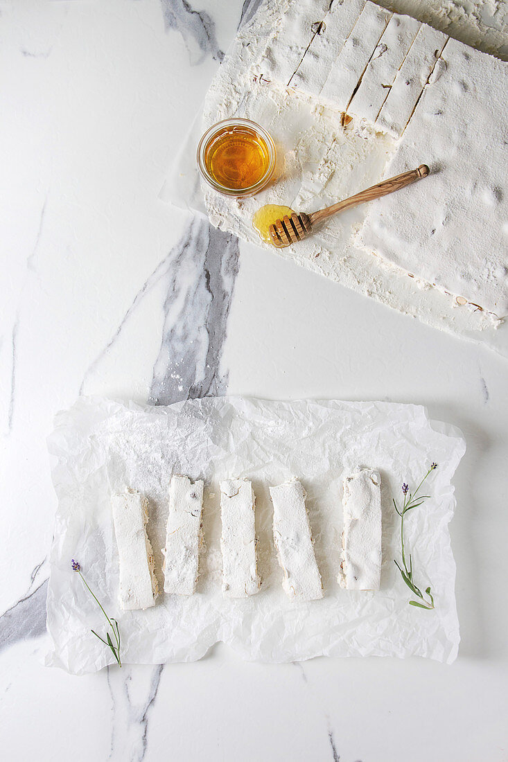 Homemade honey nuts nougat turron sliced on crumpled paper with lavender, glass of honey and sugar powder over white marble background