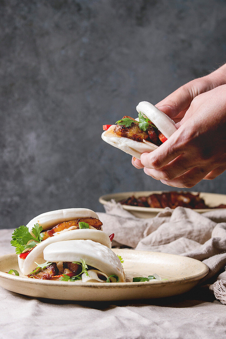 Man's hands hold asian sandwich steamed gua bao buns with pork belly, greens and vegetables served in ceramic plate