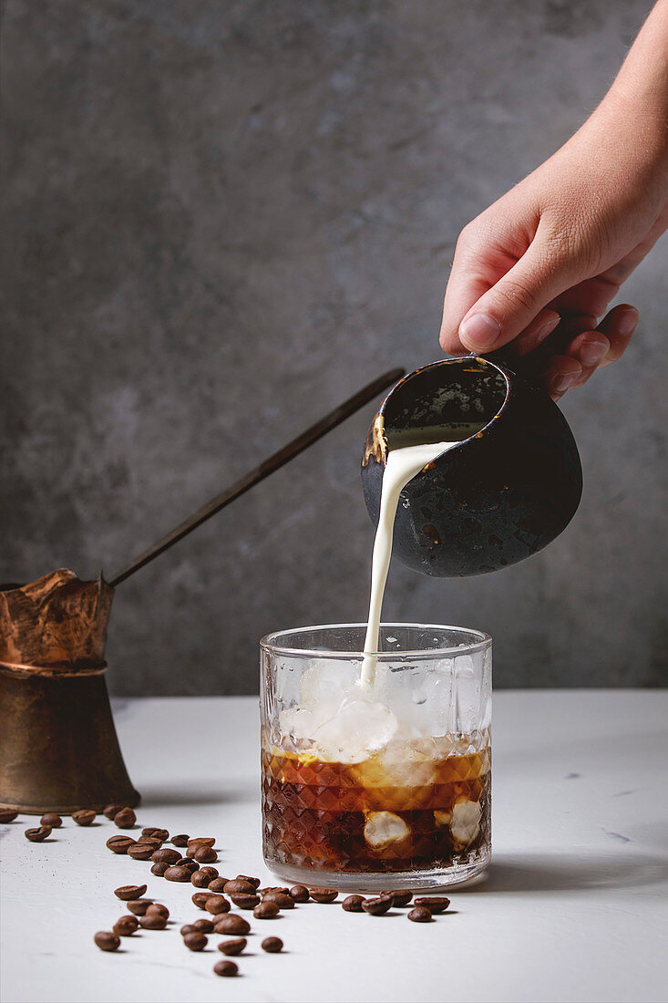 Iced coffee cocktail or frappe with ice cubes and cream, pouring from jug, served in glass with vintage jezva and coffee beans around on white marble table