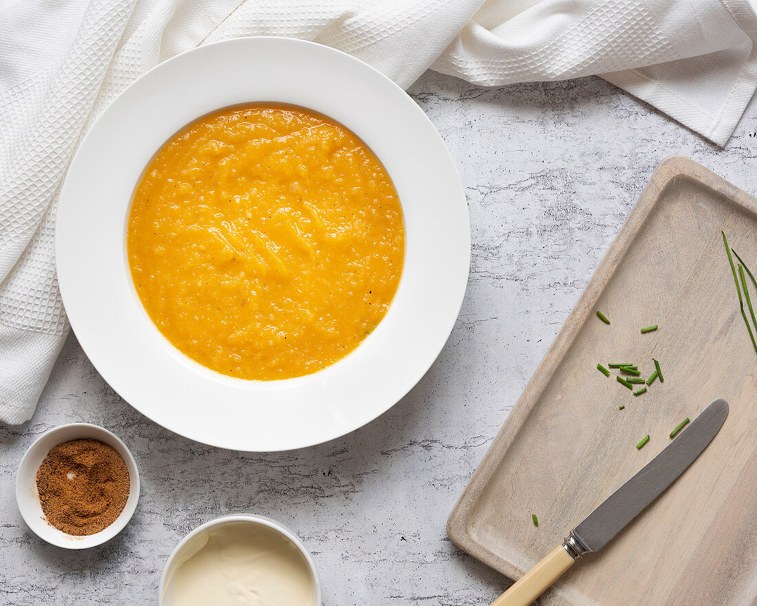 A bowl of homemade pumpkin soup beside small bowls of nutmeg and cream and chives on a wooden cutting board