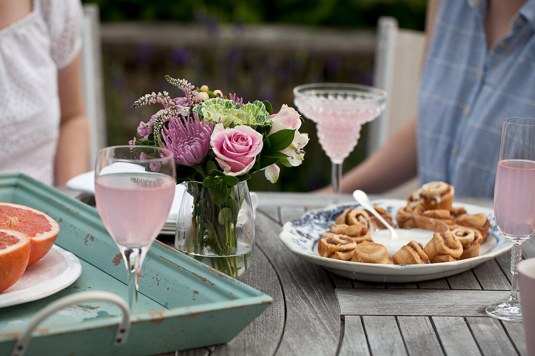Two women sitting at an outdoor brunch table with flowers, grapefruit halves, glasses of sparkling strawberry lemonade, cinnamon buns and icing