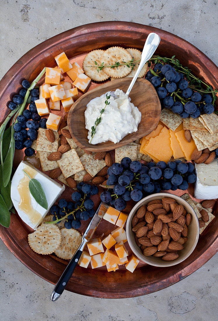A copper tray on a stone surface, with marbled cheddar cheese cubes, concord grapes, blueberries, crackers, brie topped with honey and sage, artichoke dip with thyme, almonds and a sprig of sage