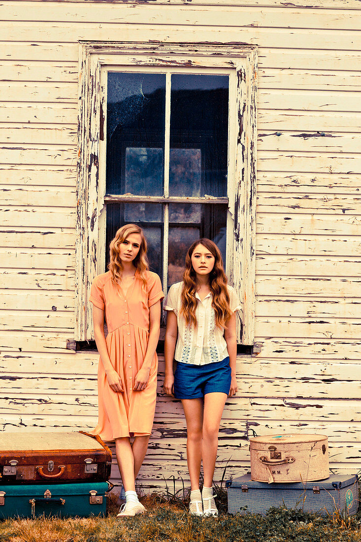 Zwei junge Frauen mit Koffern vor Holzhaus mit abgeblätterter Farbe