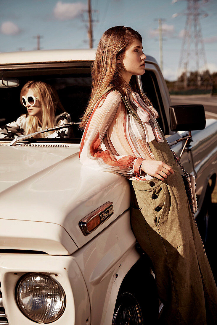 A young woman leaning against a pick-up truck and her friend wearing sunglasses sitting in the seat