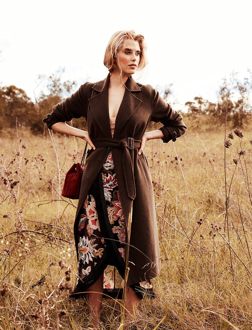 A blonde woman in a meadow wearing a patterned dress and a brown coat