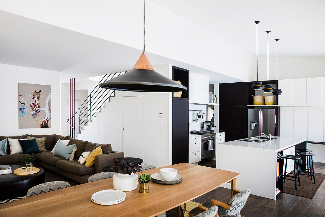 Long dining table in front of lounge with upholstered sofa, black-and-white kitchen with counter in open living room in background