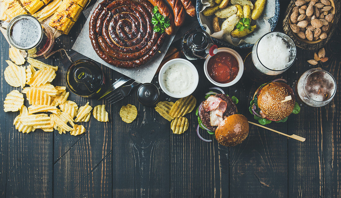 Variety of beers, grilled sausages, burgers, fried potato, corn, chips and sauces on dark wooden background