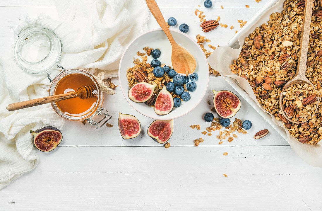 Oat granola with nuts, yogurt, honey, fresh figs and blueberries in bowl on white wooden background