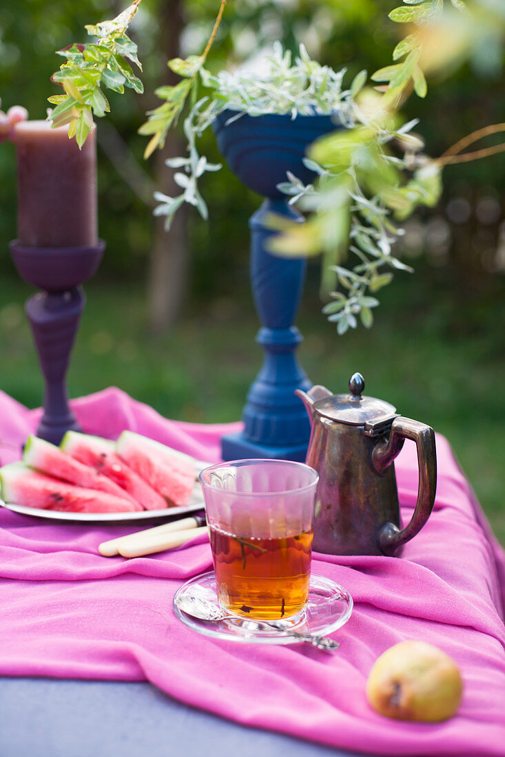 Glass of tea, melon slices and painted goblet on table in garden